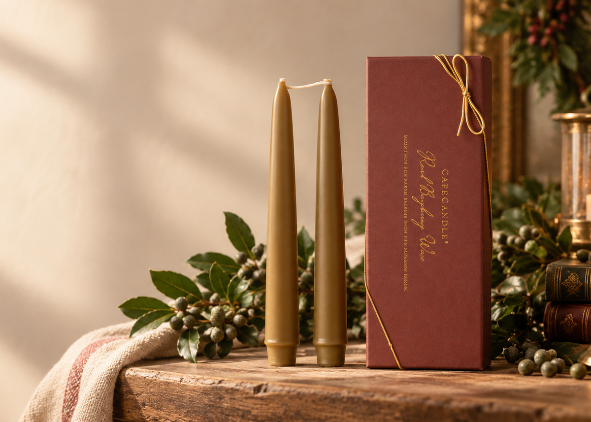 Two tall taper candles in a warm beige tone stand beside a dark red gift box with gold lettering and ribbon, styled on a rustic wooden table with holiday greenery, books, and a lantern in a softly lit interior.