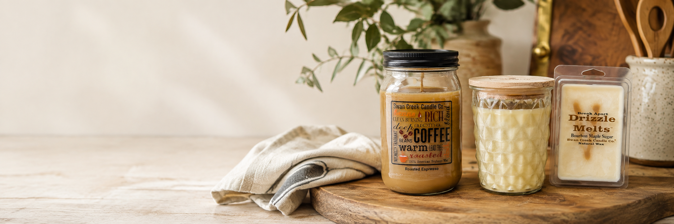 Three artisanal candles displayed on a rustic wooden board with a neutral fabric napkin. The set includes a large jar candle with a black lid and a coffee-themed label, a small glass jar with a wooden lid, and a rectangular wax melt package labeled 'Drizzle Soaps'.